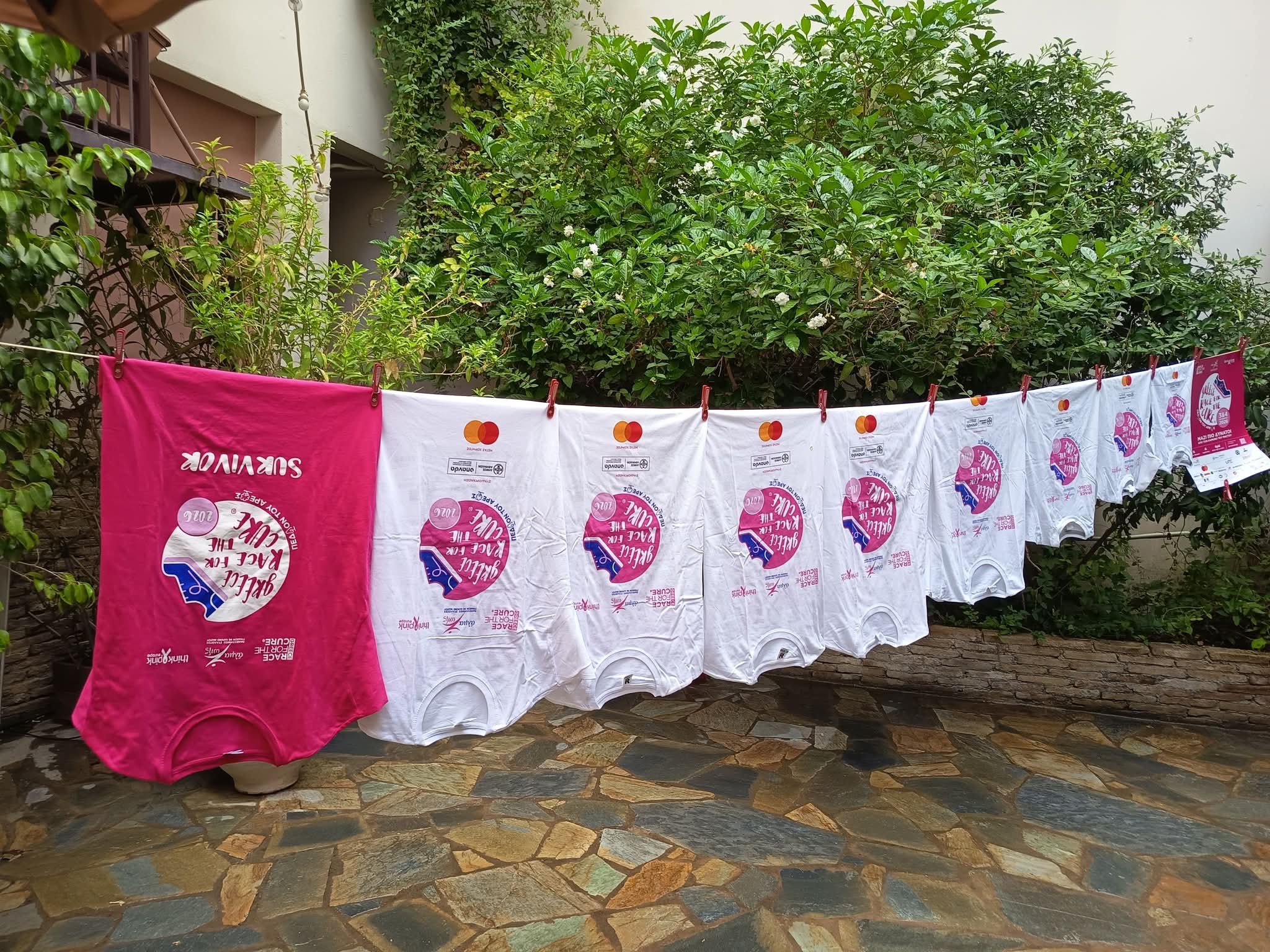 Μία ροζ και πολλές λευκές μπλούζες του Race for the Cure κρεμασμένες σε σχοινί σε μια αυλή.